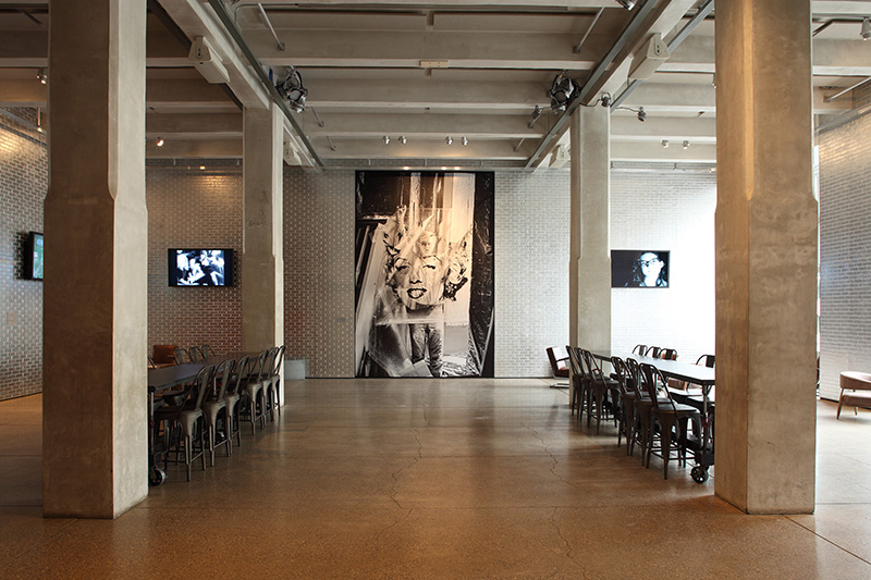 View of a large room displaying a floor to ceiling image of Andy Warhol holding a print of Marilyn Monroe's face in front of him. On each side of the Warhol image is a long table with chairs. TV screens are mounted to the walls along the perimeter of the room.