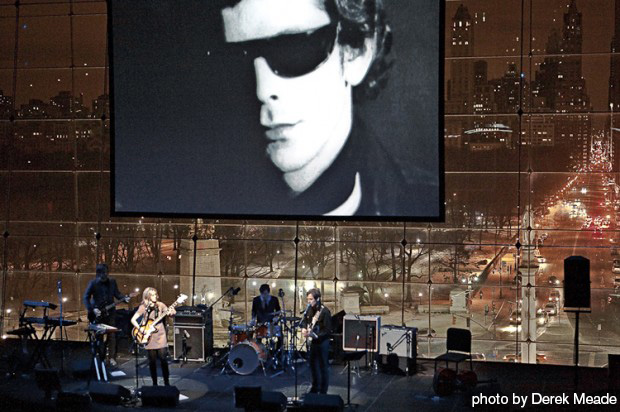 A four piece band plays in from of a glass wall through which a nighttime city-scape is visible. A large image a man wearing sunglasses hangs on the glass wall.