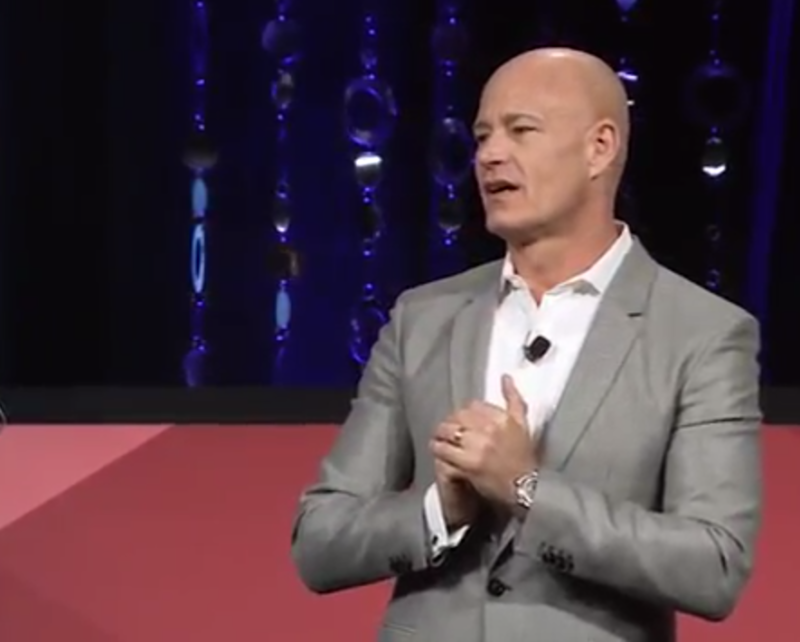 A bald man wearing a light grey suit jacket holds his hands in front of his chest. He is standing in front of a purple and pink background.