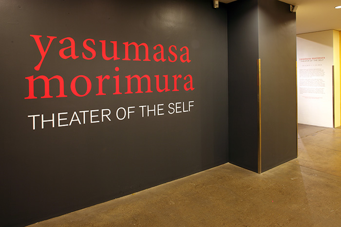 Yasumasa Morimura: Theater of the Self in red and white paint on a dark grey wall.