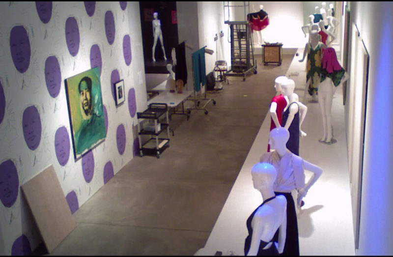 View of a hallway. On the right side of the hallway is a raised platform upon which mannequins in a variety of garments stand. There is a doorway halfway down the left side of the hall. The wall on one side of the door has multiple reproductions of a person's face painted on it in purple. A portrait of a man hangs in the middle of that wall. The wall on the other side of the door is white. Carts, ladders, and folding tables are lined up against the left side of the hallway.