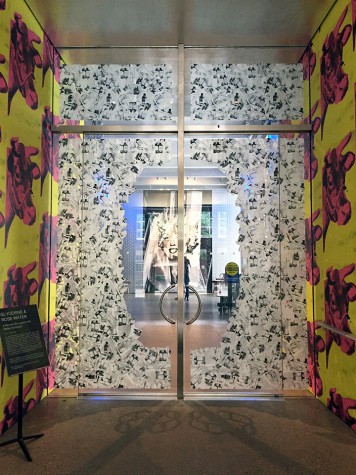 Interior view of an installation showing screen printed cows on two walls and a floral motif painted at the periphery of a glass wall looking onto a portrait of Marilyn Monroe.