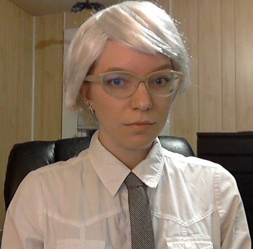 A photograph of a woman from the chest up, seated in a black leather office chair. She is dressed as Andy Warhol, wearing a white button up shirt, thin tie, glasses, and a white wig.