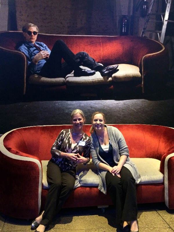 Two women sit together in the middle of a red velvet couch in the lobby of The Warhol. On the wall behind them is a large photograph of Andy Warhol reclining on the same red velvet couch.