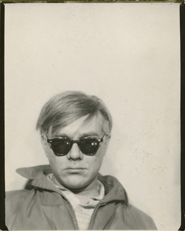 In this black and white photograph, Andy Warhol appears from the shoulders up on the left side of the image. He wears a hooded jacket and dark sunglasses.