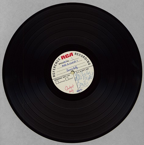 A vinyl record sits against a gray background. On the paper in its center is David Bowie's signature in blue ink.