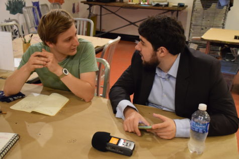 Two men are engaging in conversation, seated at a brown paper cover table in an art studio. The ma on the left has an open notebook on the table in from of him, and the man on the right is holding an iPhone.