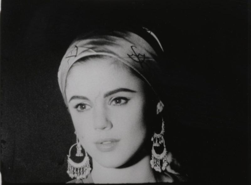 In a still from one of Warhol's a black and white screen test films, Edie Sedgwick looks towards the bottom left of the screen. Her lips are slightly parted. She wears a scarf around her hair and large, dangling earrings that stand out against the black background of the shot.