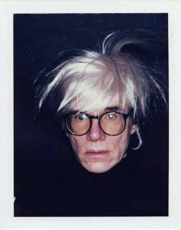 A photograph of Andy Warhol wearing large, round glasses and a black turtleneck that blends in with the black background. His white wig is tossed and hair sticks out in all directions.