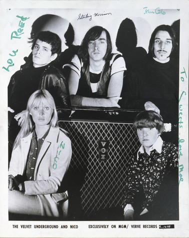 Three members of The Velvet Underground stand behind a large amp in this black and white photograph. Another band member and the artist Nico sit in front of the amp. The members have all signed the photograph near their picture in green marker or black pen.