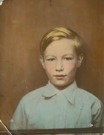 A photograph of Andy Warhol as a child. He wears a pale blue button up shirt and stands against a solid brown background. His hair is faintly gold, and there is a tint of pink to his cheeks and lips.