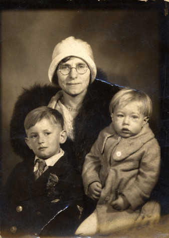 Julia Warhol, a thin woman wearing a white hat and dark fur coat, poses with two of her sons. On the left side of the image is John, a school-aged child, wearing a suit and tie. On the right, sitting on Julia's lap, is Andy Warhol, at this point a blonde haired toddler wearing a heavy grey coat.