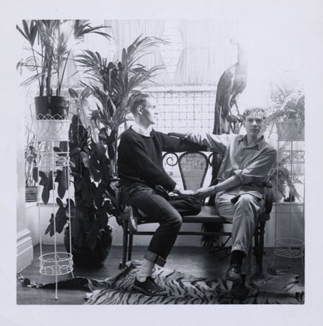 Andy Warhol as a young man seated on a bench with another person with their feet on a tiger-skin rug and houseplants in the background.
