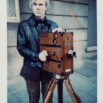 Andy Warhol poses outdoors with a silent film camera.