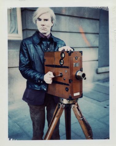Andy Warhol poses outdoors with a silent film camera.