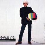 Andy Warhol stands facing front balancing a small television against his hip in a Japanese ad.