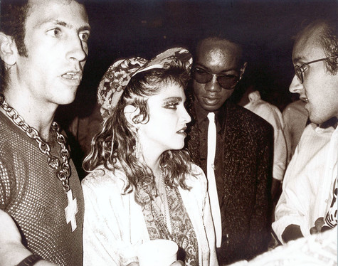 A gelatin silver print of a wide-eyed Kenny Scharf standing with Madonna, who is wearing heavy eye makeup and a printed cap. Juan Debose stands slightly behind them wearing dark sun glasses, a dark suit, and a white tie, and Keith Haring's glasses-adorned face is also visible in profile on the right side of the image.
