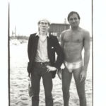 Andy Warhol, dressed in a blazer, shirt, and trousers, stands on a beach next to a person wearing a speedo style swimsuit.