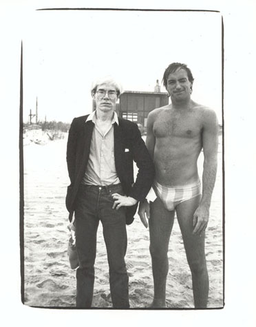 Andy Warhol, dressed in a blazer, shirt, and trousers, stands on a beach next to a person wearing a speedo style swimsuit.