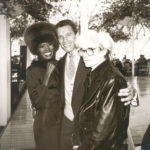 Grace Jones, Arnold Schwarzenegger, and Andy Warhol stand in an outdoor pavilion in front of an empty dance floor.