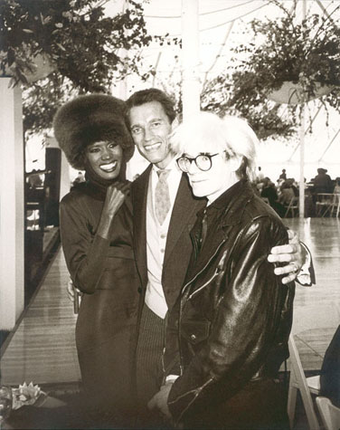 Grace Jones, Arnold Schwarzenegger, and Andy Warhol stand in an outdoor pavilion in front of an empty dance floor.