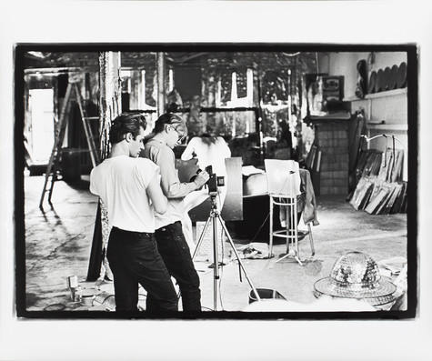 A black and white photograph of Andy Warhol in his studio, wearing a long-sleeved shirt and black pants, adjusting a camera on a tripod. Another man wearing a white t-shirt and black pants observes.