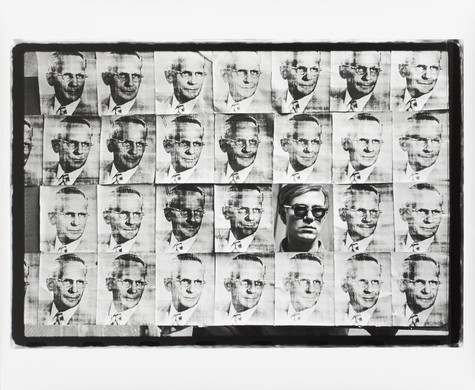 A black and white photograph of Andy Warhol wearing dark sunglasses peeking through his American Man piece, which features a grid of images of a man with short hair in a suit.