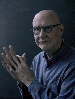 A bald man wearing glasses and a long sleeved blue shirt looks at the viewer with his hands loosely clasped in front of his chest.