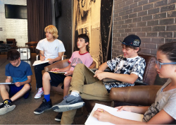 Five young people holding sketchpads sit in a row. Four sit in chairs and one sits on the floor.