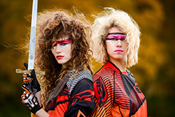 Two long-haired people wearing bright multicolored clothing and face paint stand back to back. The person on the left holds up a sword in both hands with the blade in front of their face.
