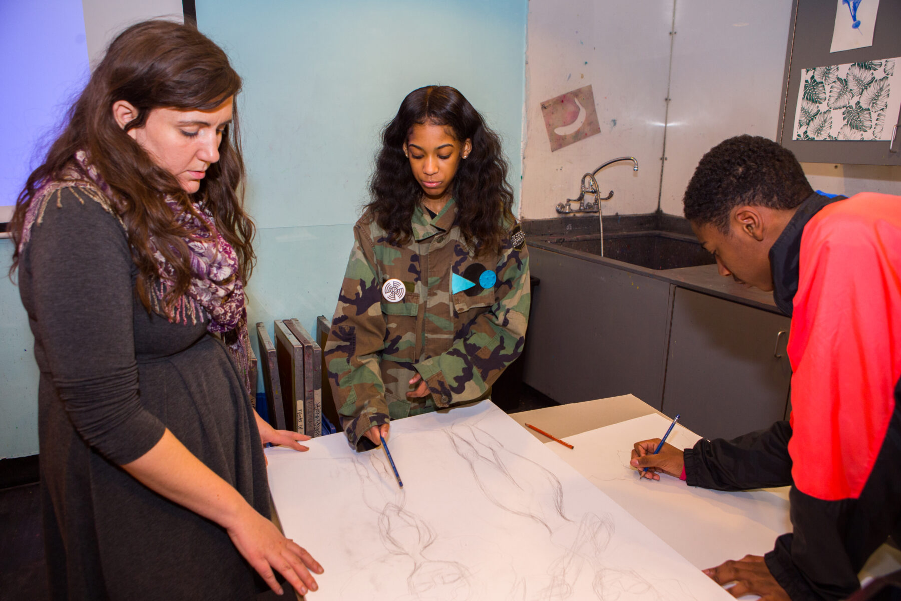 Three individuals examine sketches of an exaggerated female figure on a table in The Factory.