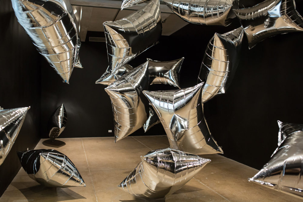 The photograph depicts an installation piece at the Warhol museum. The floor is tan, and the walls are black, and large, rectangular, metallic silver balloons fill the space, drifting chaotically between the floor and ceiling.