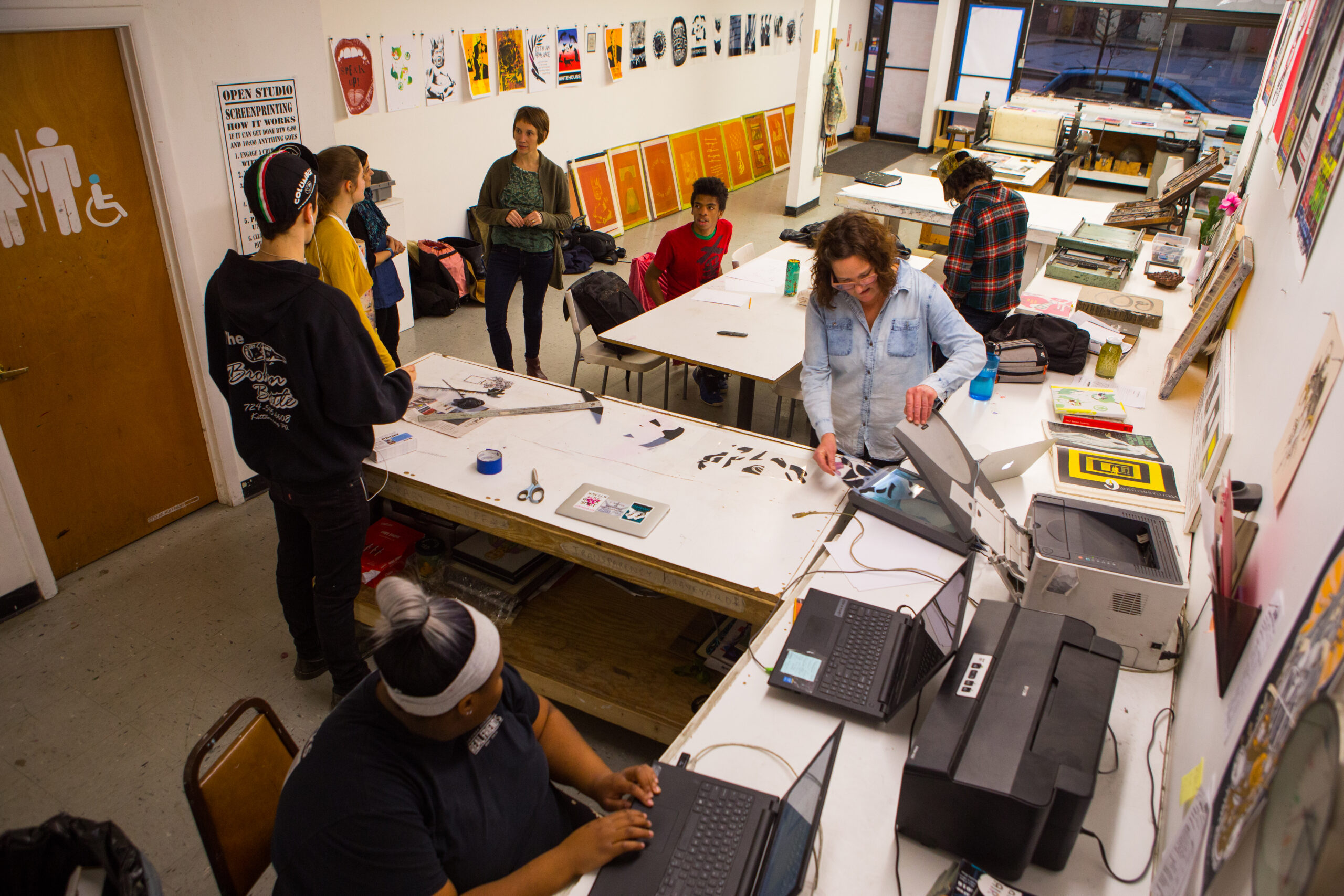 A studio full of young people working on various phases of the printing process. A young woman types on a laptop, a woman arranges papers in a scanner, and several teens are gathered around counters and tables in the room talking.