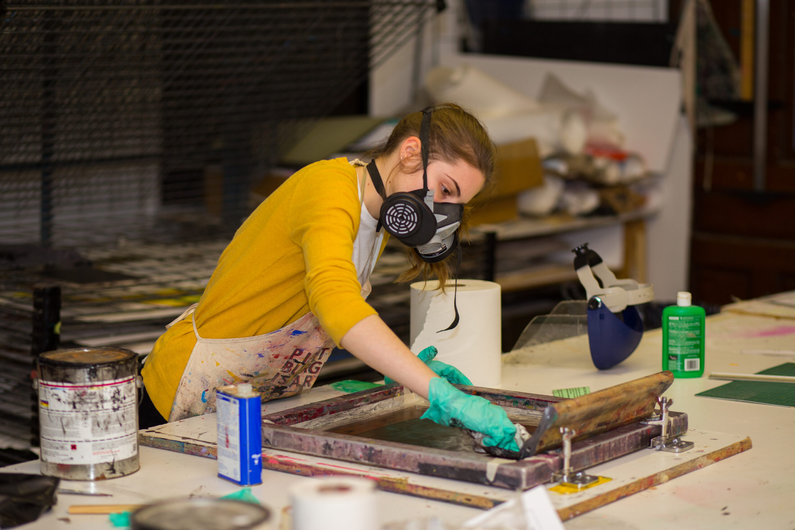 A young woman in a yellow cardigan wears green gloves and a mask as she prepares a screen for printing.