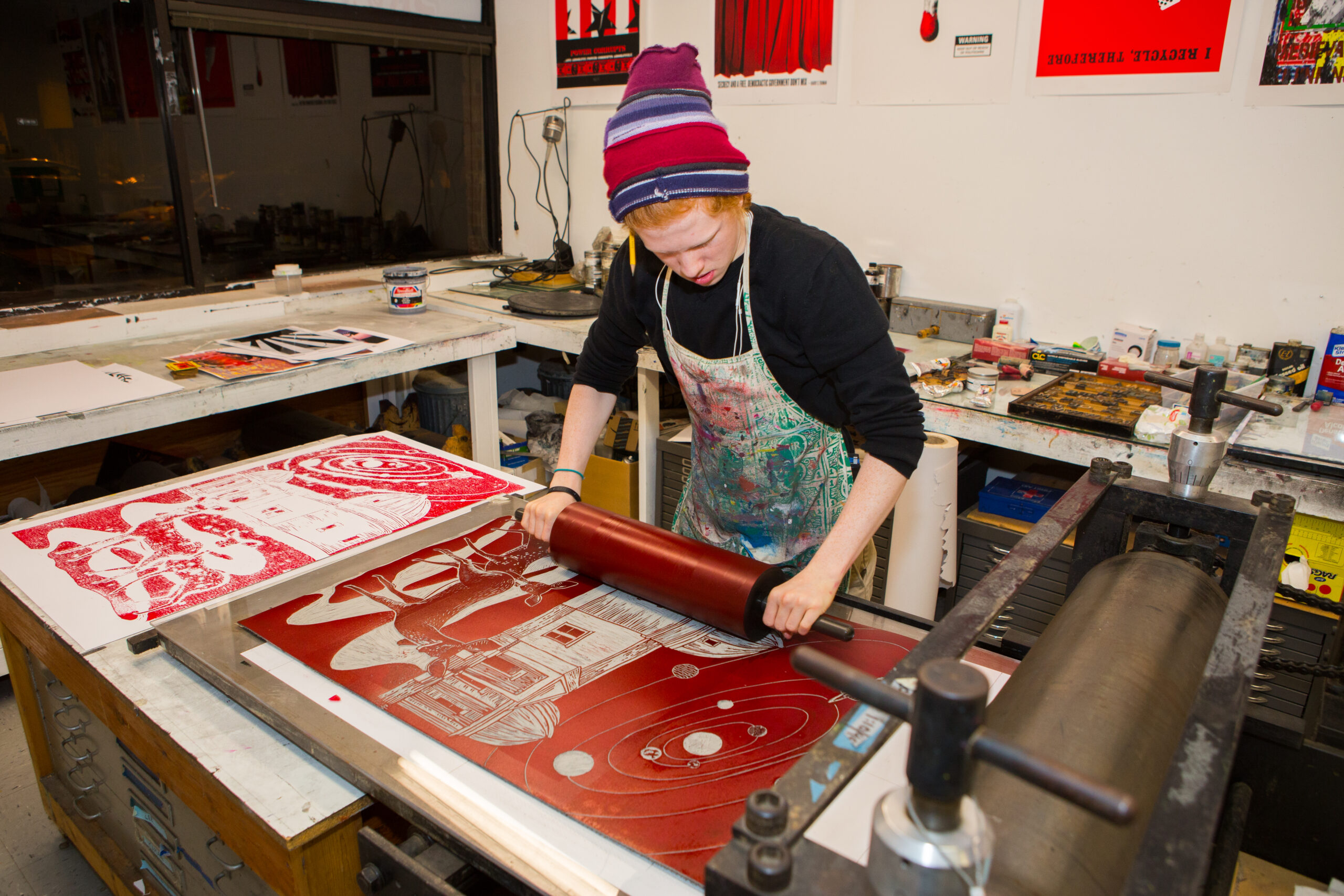 A young man with red hair wearing a red, white, and blue beanie, a black sweatshirt with the sleeves rolled up, and a paint-splattered apron rolls ink onto a carved linoleum block for printing.