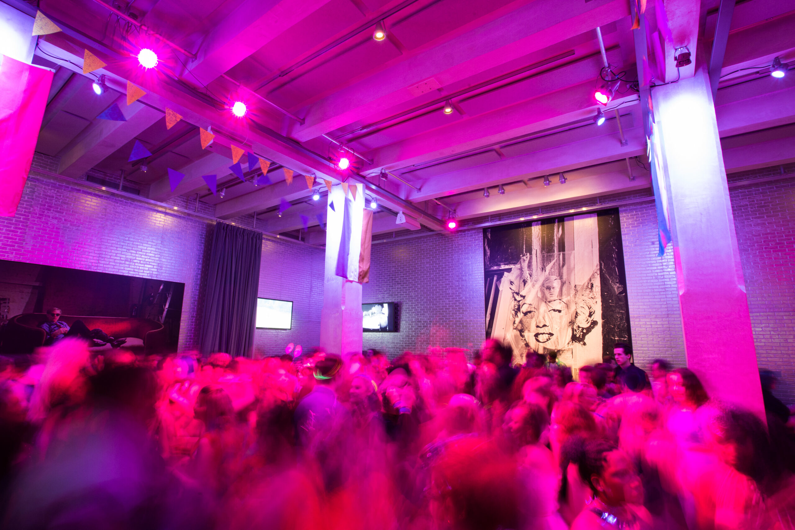 A large group of teenagers are in the lobby of The Andy Warhol Museum dancing. They are blurry due to their movement. There is pink lighting throughout the image. An image of Marilyn Monroe with Andy Warhol superimposed within it is in the background and to the left there is a photograph of Andy Warhol laying on a red couch on the wall. There are also two small video screens in the background hanging on the wall.