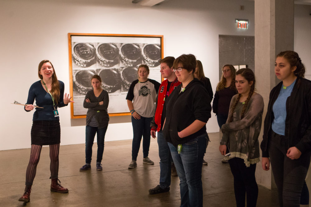 A woman with long brown hair in a black dress who is wearing an Andy Warhol Museum staff badge gestures as she leads a group of students in an in-gallery discussion.