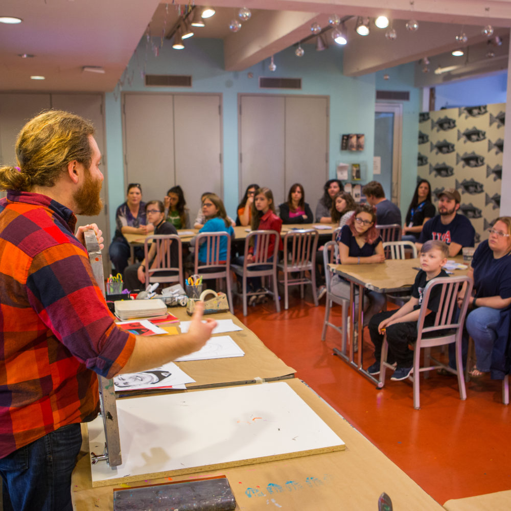 A male artist educator with long blond hair pulled back into a bun wearing a red plaid shirt presents information to students sitting in silver chairs at long, rectangular tables in the Andy Warhol Museum’s studio.