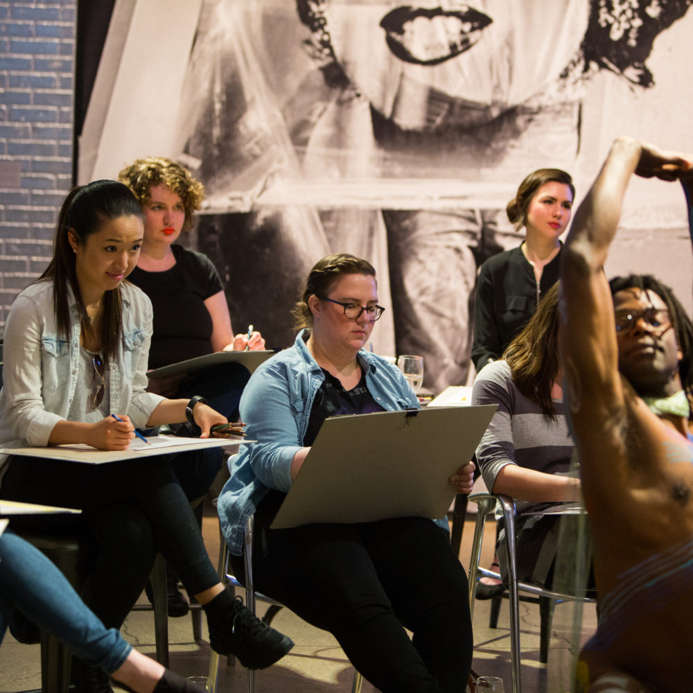 Sip and Sketch participants sit around an African American male nude model who is posing, arms overhead, in body paint during an event in the Andy Warhol Museum entrance space.