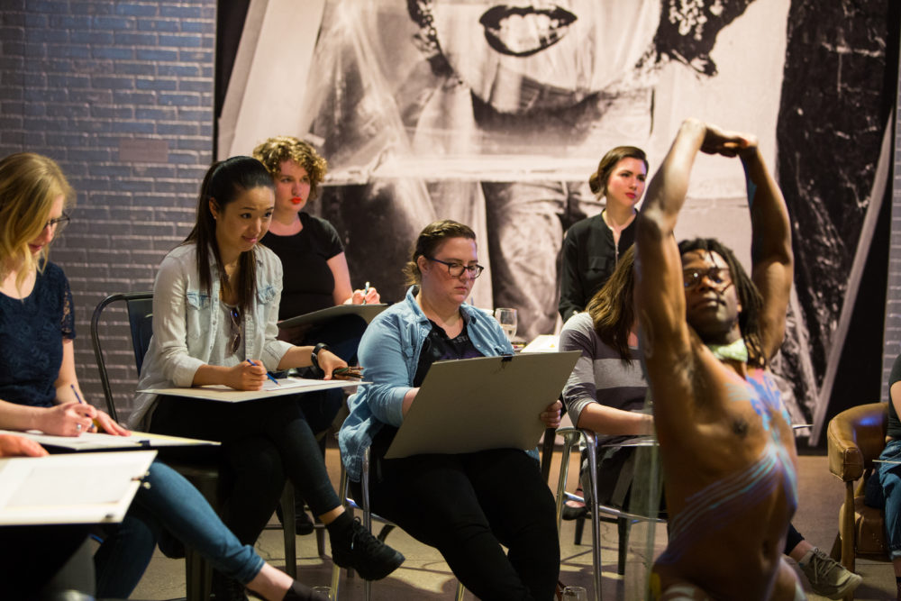 Sip and Sketch participants sit around an African American male nude model who is posing, arms overhead, in body paint during an event in the Andy Warhol Museum entrance space.