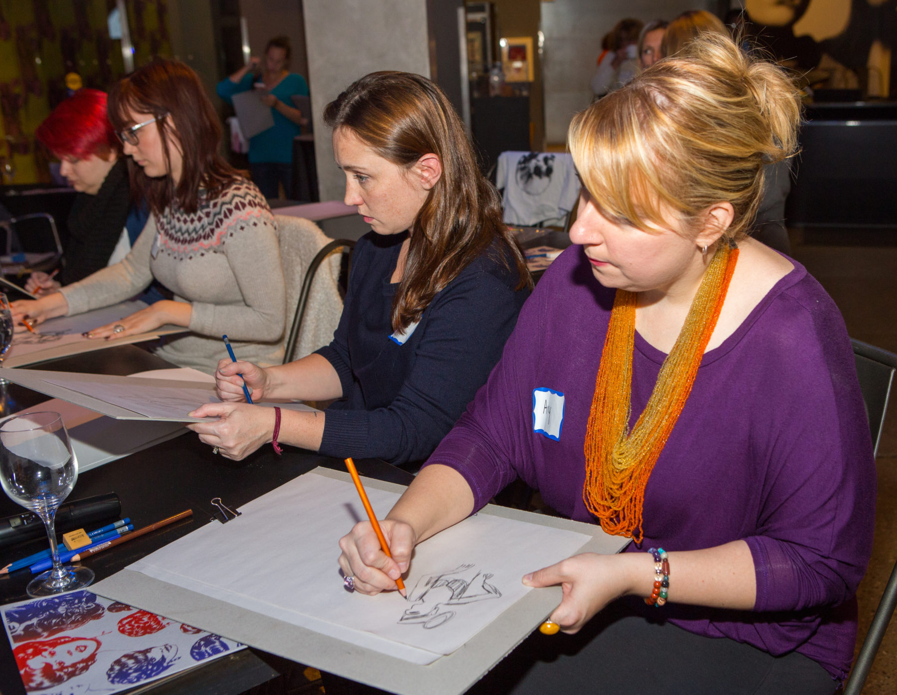 Four women sit at a long table sketching with pencil.