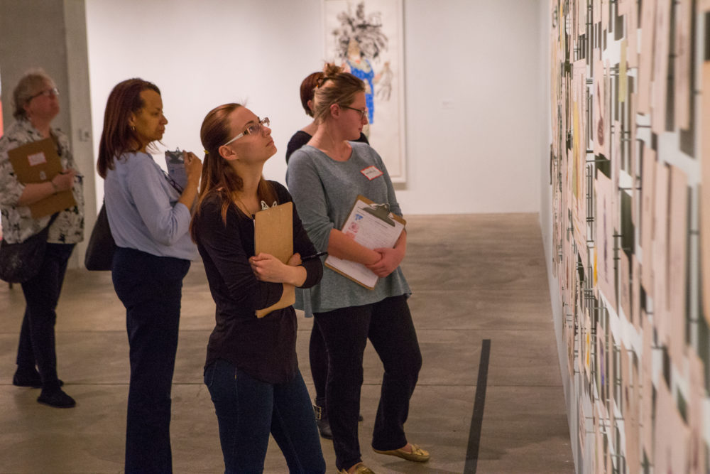 Five women look at a wall of numerous small drawings hung salon style.