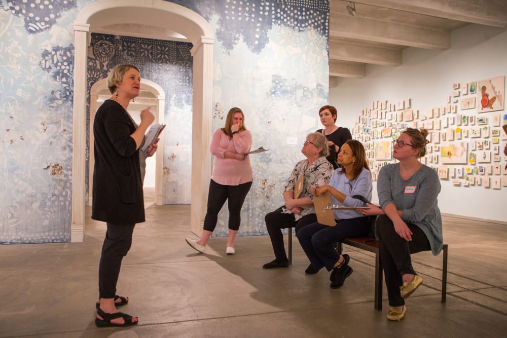 A woman with short blonde hair speaks to a group of colleagues holding clipboards and sitting on a bench in the Andy Warhol Museum's 2nd floor gallery during the Firelai Baez Exhibition.