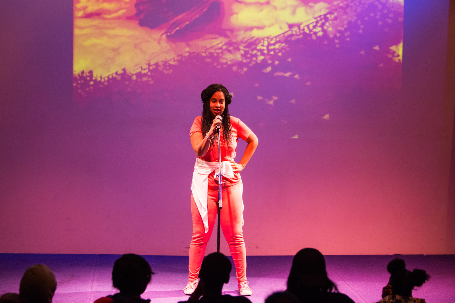 A female African American student holds a microphone stand in one hand and places her other on her hip as she performs against a background illuminated with pink and purple light.