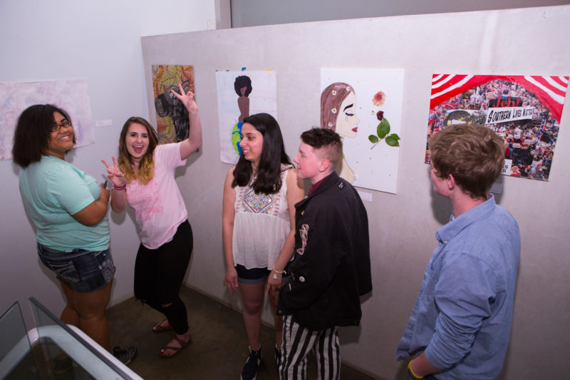 Five teenagers standing in a space with white walls. There are three girls on the left side and two boys on the right. The second girl from the left is making the peace sign with both of her hands. Behind the teenagers are five artworks that were made by teenagers.