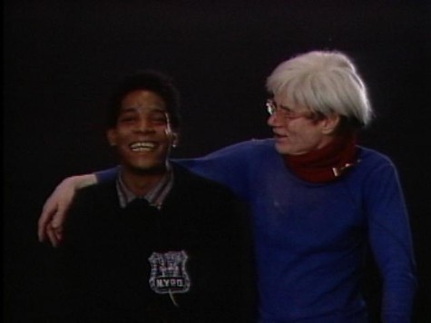 White-haired Andy Warhol, wearing glasses and a purple sweater over a burgundy turtleneck, places his arm around young African-American boy wearing and NYPD badge.