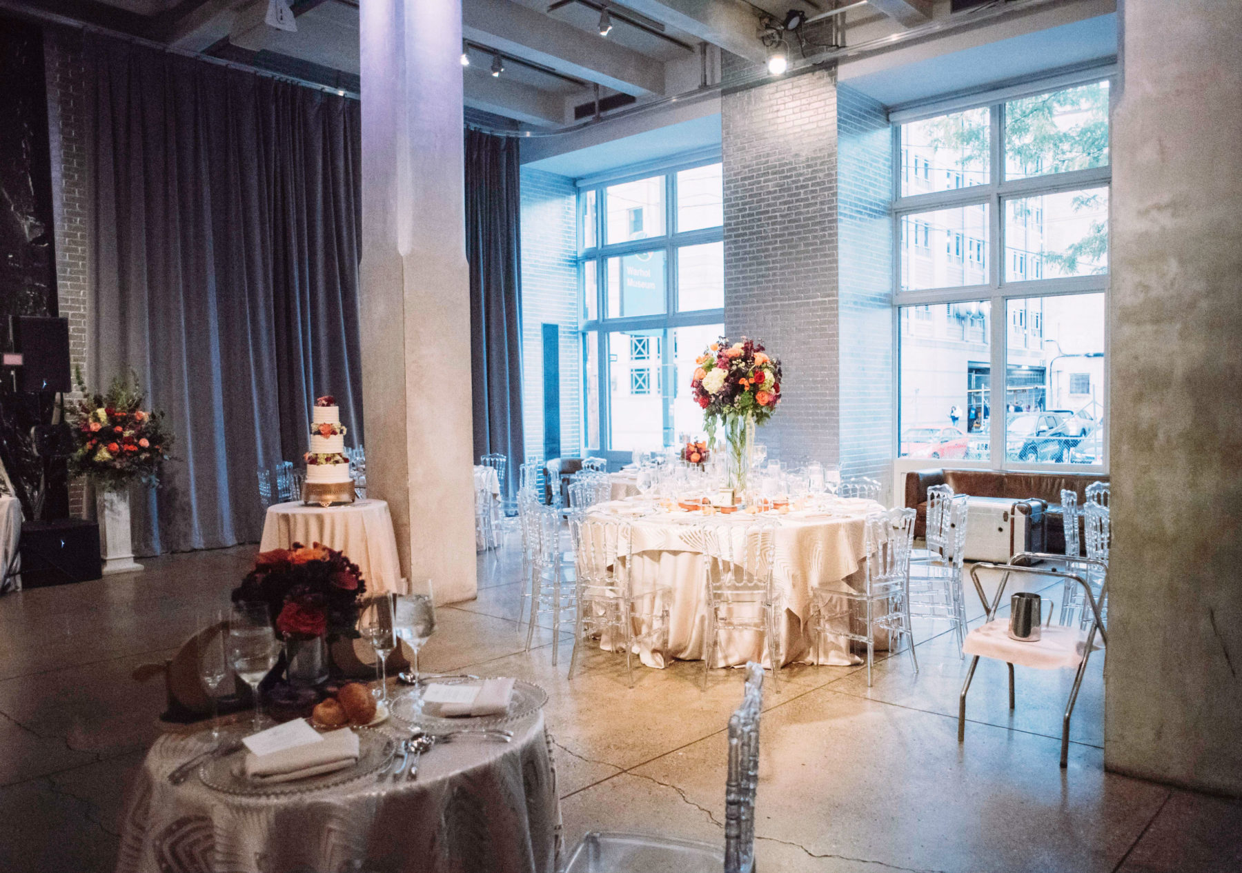 The Warhol entrance space with silver brick walls, gray curtains, two silver pillars, and large windows in the background. Tables with white tablecloths and clear, plastic chairs and flower centerpieces fill the lobby. A tiered wedding cake is on a table in the back left of the photo with a sweetheart table near the front of the photo.