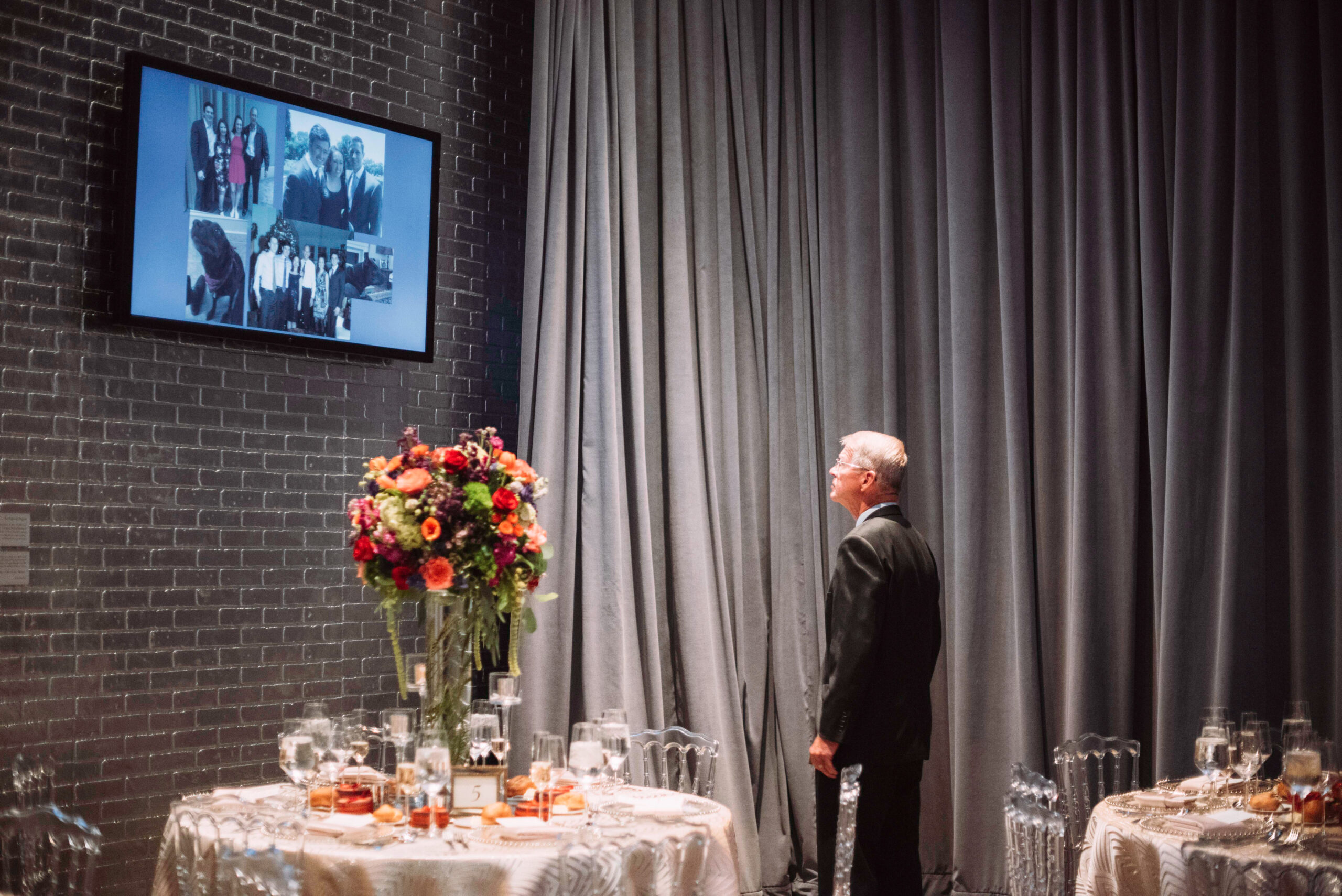Man in a suit looks up at screen with photographs on it. In the foreground is a table with white linen, clear plastic chairs, and a flower centerpiece. Gray curtains and a silver chair are in the background.