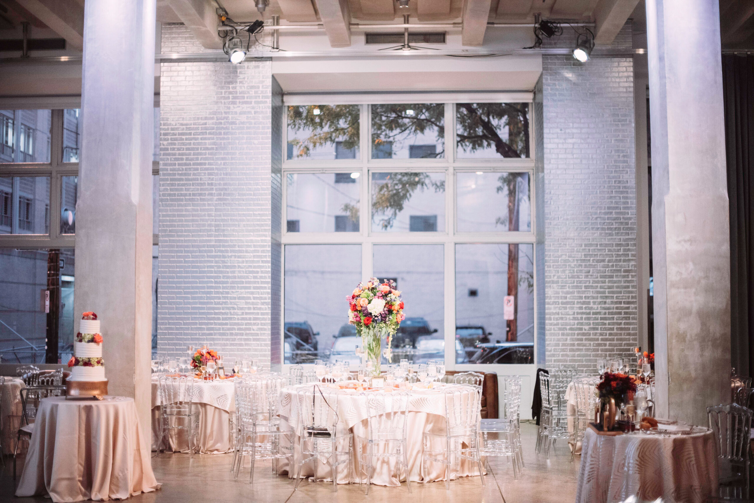The Warhol entrance space with silver brick walls, two silver pillars, and a large window in the background. Five tables with white tablecloths and clear, plastic chairs and flower centerpieces. A tiered wedding cake is on a table on the left of the photo.