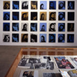 A grid of canvases in shades of blue and gold hanging on a wall. The repeated image is of Jackie Kennedy, wife of former President John F. Kennedy. In the foreground is a vitrine displaying various magazine and newspaper clippings surround the JFK assassination.
